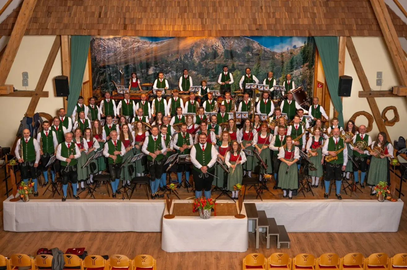 Ein großes Musikensemble, gekleidet in traditionelle grüne und weiße Uniformen, tritt auf einer Bühne mit einem Gebirgshintergrund auf. Die Musiker spielen verschiedene Instrumente, darunter Saxophone und Flöten.