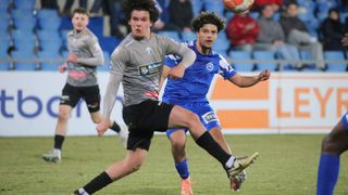 Zwei Fußballspieler kämpfen um den Ball, einer in Blau und einer in Grau, mit einem dritten Spieler, der dahinter läuft. Zuschauer beobachten vom Zuschauerrang aus.