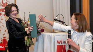 Zwei Frauen feiern mit einem Blumenstrauß und einem Zertifikat. Eine hält ein Mikrofon, die andere ein Zertifikat, an einem Tisch mit einem roten und weißen Schild.