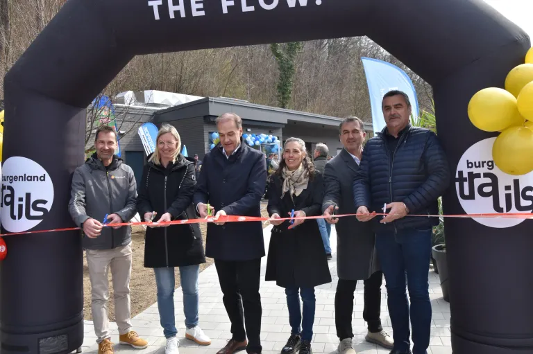 Fünf Menschen stehen in einer Reihe und halten ein Band mit der Aufschrift The Flow über ihnen. Sie alle lächeln und einige halten Scheren. Hinter ihnen stehen Leute und ein Gebäude.