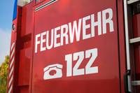 Ein roter Feuerwehrwagen zeigt das Wort 'FEUERWEHR' und die Nummer '122' auf seiner Seite, mit einem Anrufsymbol. Der Wagen ist draußen geparkt.