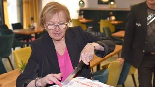 Eine ältere Frau mit Brille und einem rosa Oberteil schneidet in einem Restaurant einen Kuchen mit der Aufschrift 'Glückliche Rente'.