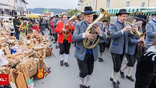 Eine Gruppe von Musikern in traditioneller Kleidung spielt auf der Straße Instrumente, während eine Menge zuschaut und Geschäfte Waren im Hintergrund verkaufen.