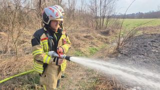 Zwei Feuerwehrleute in gelben Uniformen sprühen Wasser aus einem Schlauch auf ein Feld.