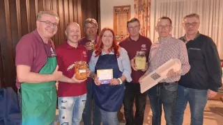 Sechs Erwachsene stehen lächelnd in einem Raum, zwei von ihnen halten Gläser mit Essen. Die Frau in der Mitte trägt eine Schürze und hält ein Zertifikat. Der Mann auf der rechten Seite hält eine Box.