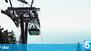 Zwei Personen in einer Seilbahn, möglicherweise einer Gondel, fahren mit Blick auf die Stadt in der Luft.