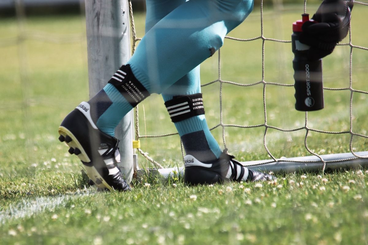 Nahaufnahme der Beine eines Fußballspielers in blauen Leggings, schwarzen Schuhen und Socken mit schwarzen und weißen Streifen. Der Spieler steht vor einem Fußballtor.