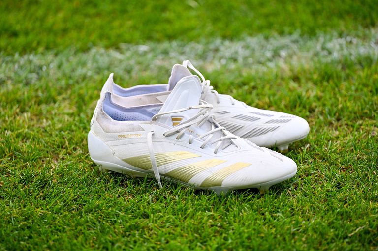 Ein Paar weiße Fußballschuhe mit gelben Streifen liegt auf dem Rasen. Die Schuhe haben ein goldenes Logo und den Schriftzug 'PREDATOR' an der Seite.