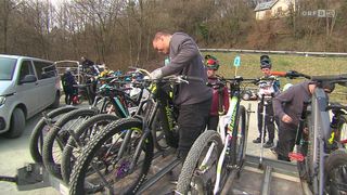 Ein Mann inspiziert ein Fahrrad vor einem Rennen in einem bewaldeten Gebiet mit einem Haus und Bäumen im Hintergrund.