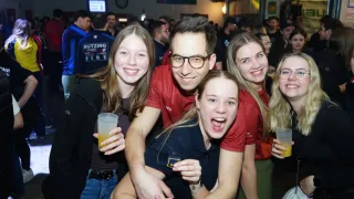 Fünf Menschen lächeln in einer Party-Atmosphäre zur Kamera. Ein Mann mit Brille hat seine Arme um zwei Frauen. Die Frau auf der linken Seite hält ein Glas mit einem Getränk.
