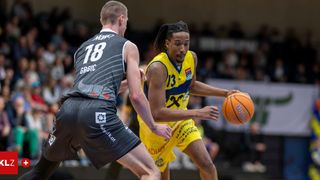 Zwei Basketballspieler in Aktion. Einer in Gelb mit der Nummer 13, der ballführt, der andere in Grau mit der Nummer 18, der verteidigt. Sie spielen in einer überdachten Arena mit einem jubelnden Publikum.