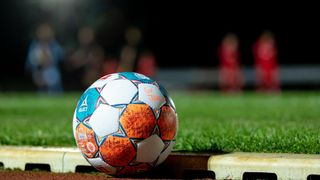 Ein Fußball mit blauen, weißen und orangefarbenen Mustern steht auf einem Fußballfeld. Im Hintergrund sind verschwommene Menschen in Rot zu sehen.