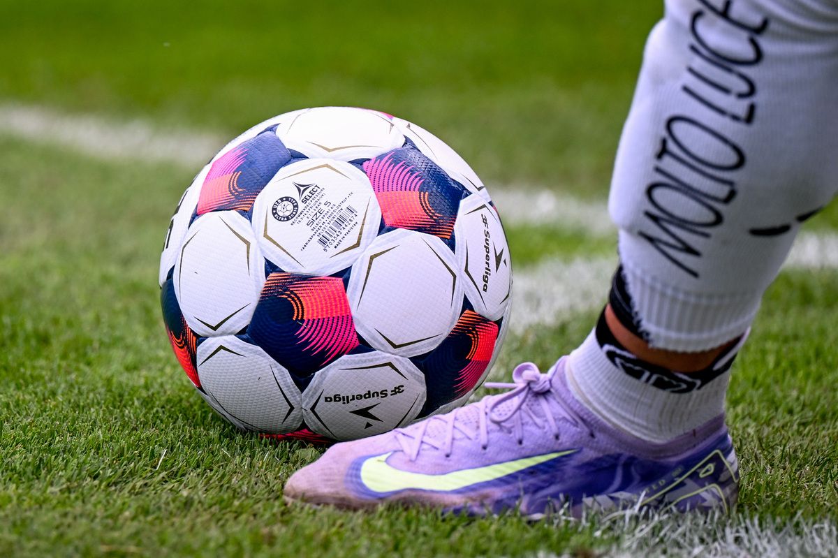 Ein Fußball mit der Aufschrift 'Select' in leuchtenden Farben liegt auf dem Feld neben einem lila-weißen Fußballschuh.