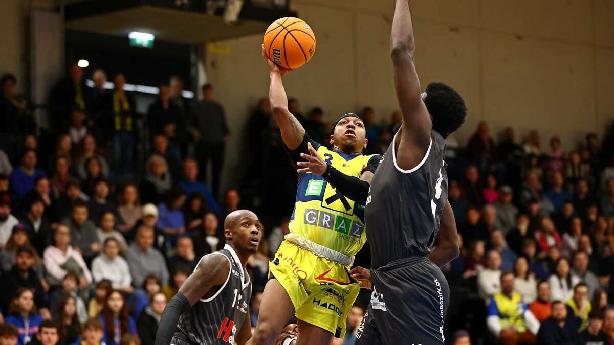 Basketballspieler treten während eines Spiels gegeneinander an. Ein Spieler in einem gelben Trikot springt zum Werfen, während ein anderer in Schwarz versucht, zu blocken. Zuschauer schauen von den Tribünen zu.