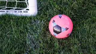 Ein rosafarbener Fußball mit blauen Diamantmustern liegt auf einem Rasenfeld neben einem weißen Torpfosten.