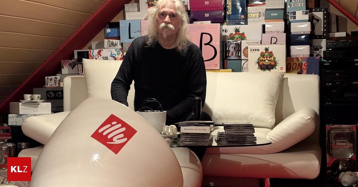 Ein älterer Mann mit weißem Bart und Schnurrbart sitzt auf einem Sofa in einem Raum voller Kartons, ein weißes Kissen an seiner Seite. Ein Tisch mit Stapeln von CDs steht vor ihm.