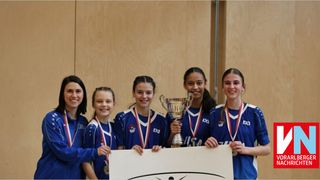 Eine Gruppe von Sportlerinnen hält eine Trophäe und Medaillen und posiert für ein Foto. Sie tragen blaue Uniformen und lächeln. Dahinter befindet sich eine Holzwand.