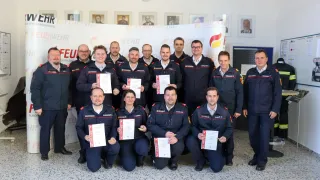 Eine Gruppe uniformierter Feuerwehrleute posiert für ein Foto, einige halten Urkunden in der Hand, in einem Raum mit weißen Wänden und gerahmten Porträts.