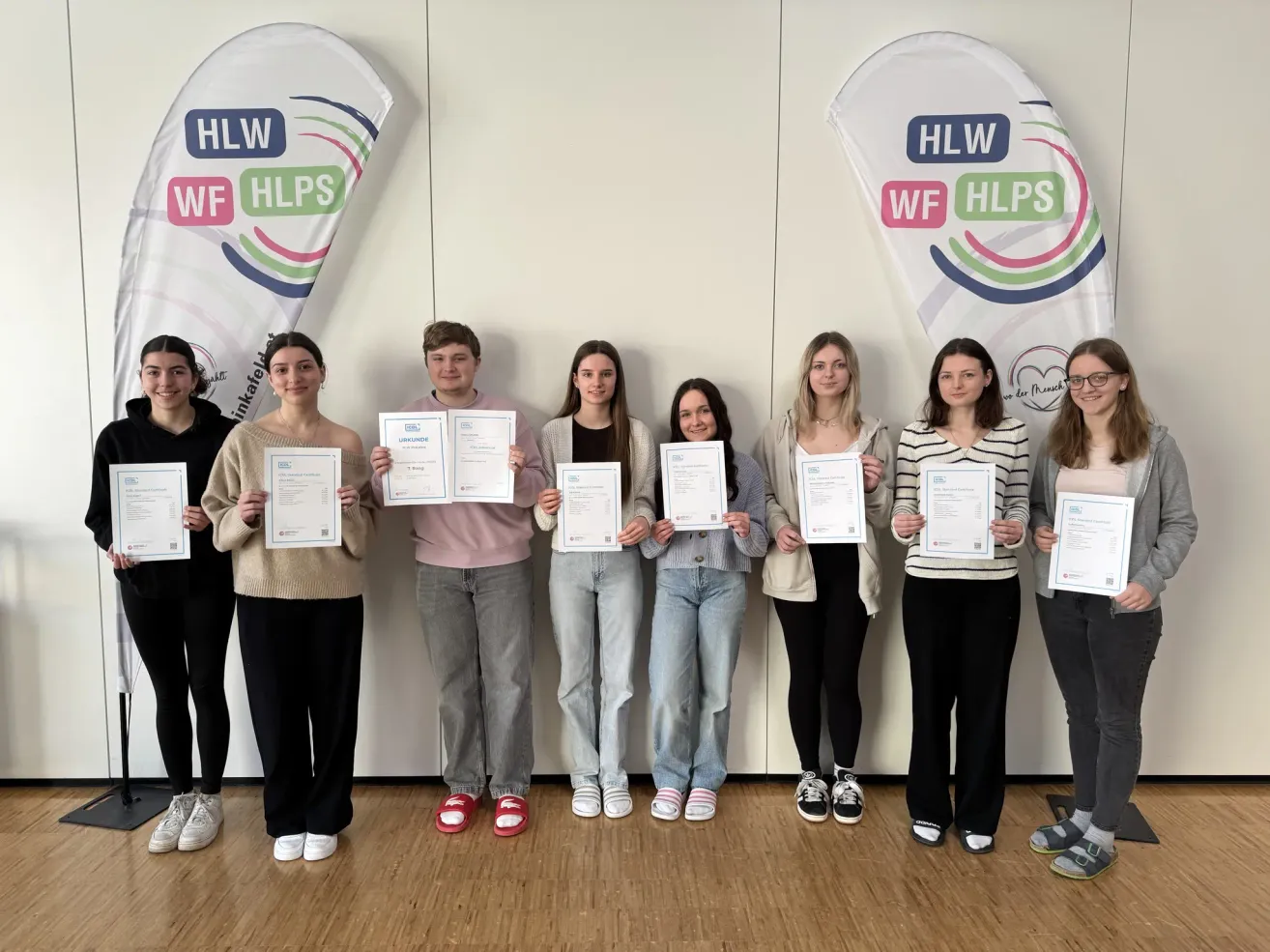 Eine Gruppe junger Erwachsener steht in einer Reihe und hält Urkunden in den Händen. Sie lächeln für die Kamera. Sie stehen vor einer weißen Wand mit Bannern mit Logos.