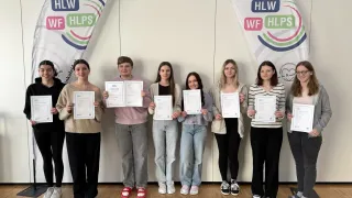 Eine Gruppe junger Erwachsener steht in einer Reihe und hält Urkunden in den Händen. Sie lächeln für die Kamera. Sie stehen vor einer weißen Wand mit Bannern mit Logos.