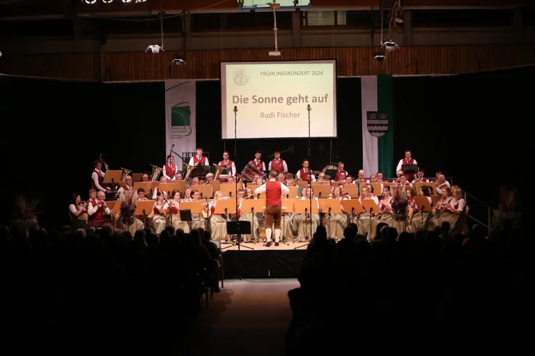 Ein Orchester spielt auf der Bühne beim Fruhlingskonzert 2026. Die Bühne ist mit Musikern gefüllt, die verschiedene Instrumente spielen. Eine große Leinwand zeigt den Titel der Aufführung.
