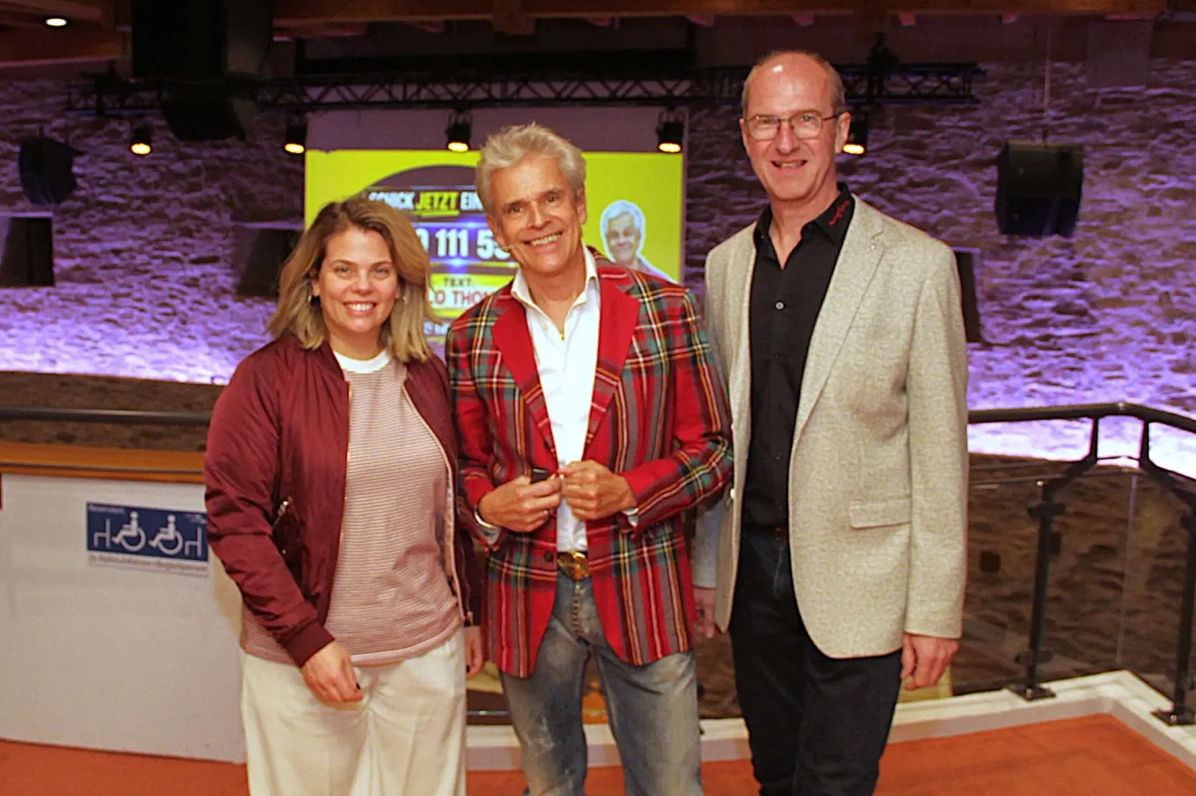Drei Menschen stehen zusammen und lächeln für ein Foto. Eine Frau und zwei Männer, einer in einer karierten Jacke und der andere in einem Anzug, mit einem gelben Banner hinter ihnen.