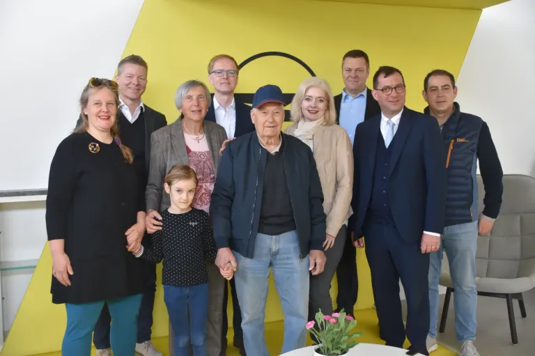 Eine Gruppe von Menschen, darunter ein älterer Mann, steht vor einem gelben Hintergrund und lächelt für ein Foto.