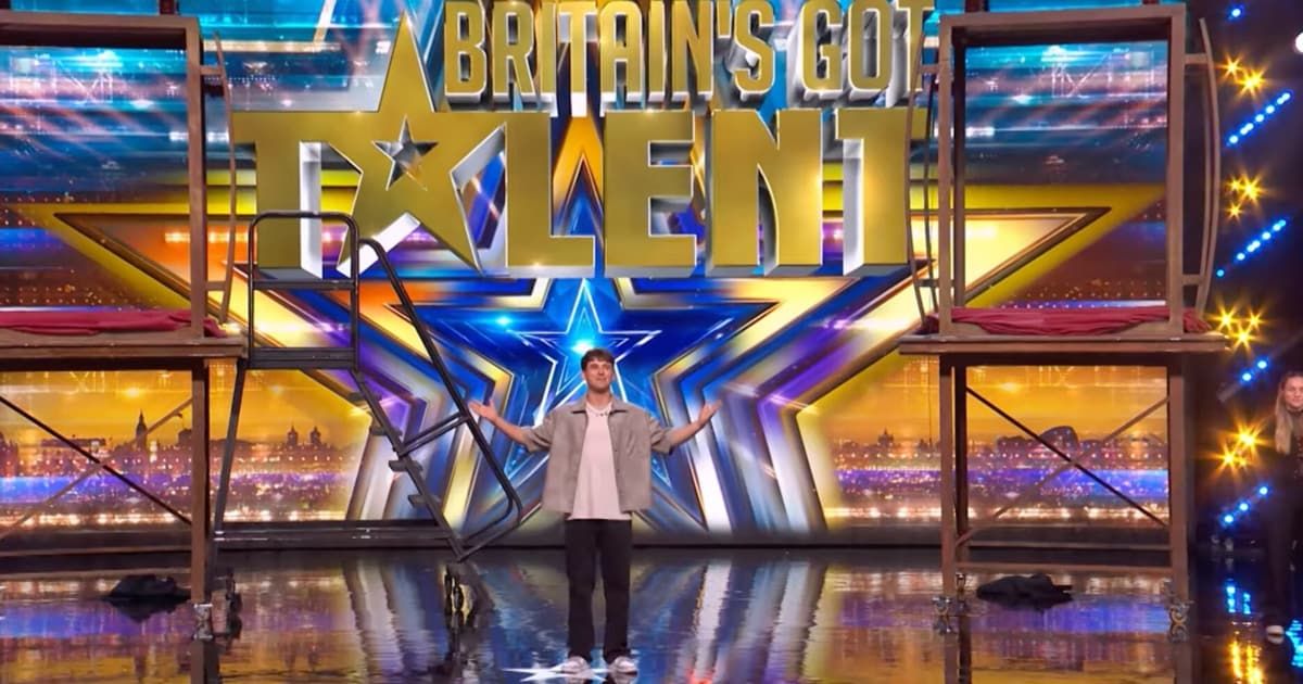 Ein Mann steht mit ausgebreiteten Armen auf der Bühne vor einer Hintergrundkulisse, die 'Britain's Got Talent' in großen Buchstaben zeigt. Die Bühne ist glänzend und auf beiden Seiten befinden sich zwei Metallkonstruktionen.