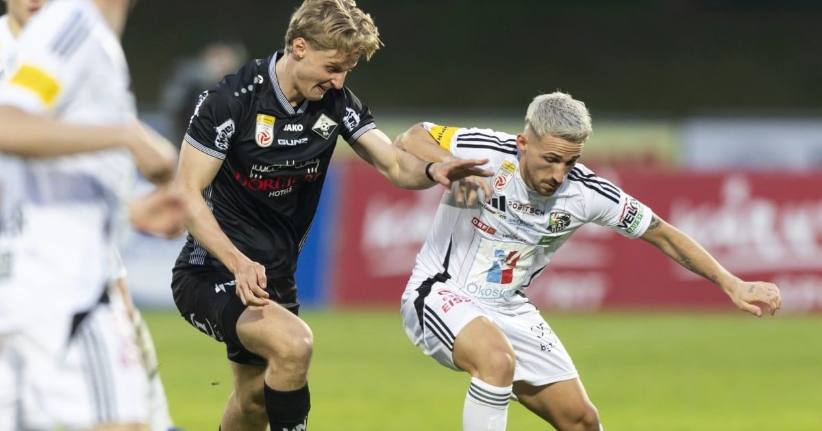 Zwei Fußballspieler, einer in Schwarz und der andere in Weiß, kämpfen auf einem grünen Feld um den Ball.