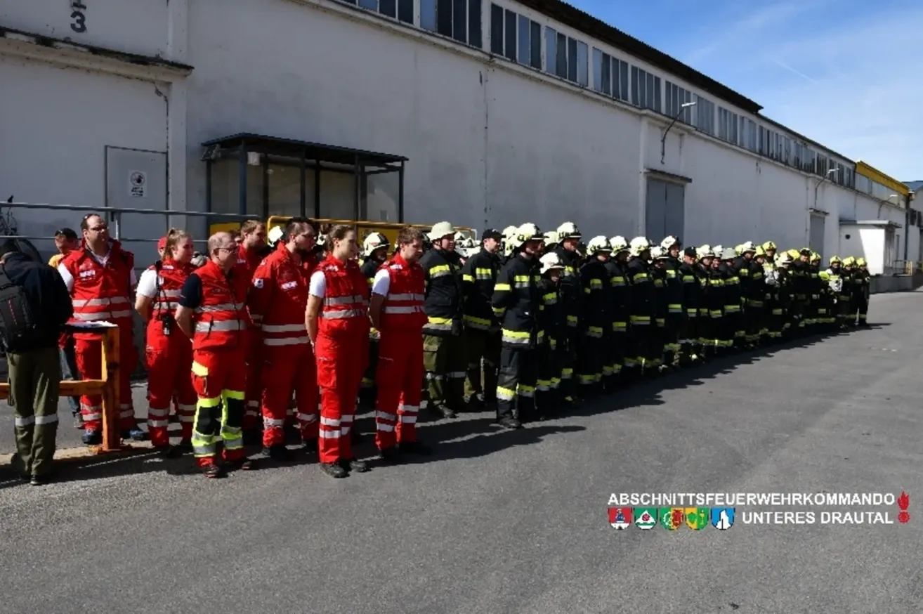 Eine Reihe von Feuerwehrleuten in roten und schwarzen Uniformen, einige tragen Helme, stehen in Formation. Hinter ihnen ein weißes Gebäude mit vielen Fenstern. Das Bild ist mit 'ASCHNITTSEUERW' und 'UNT' beschriftet.