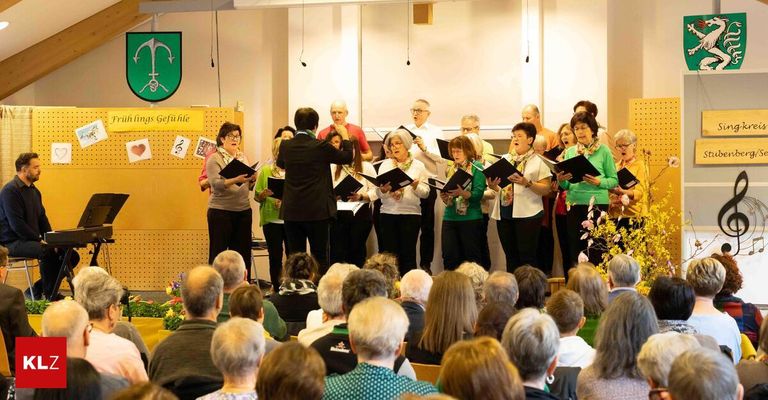Eine Gruppe älterer Erwachsener, gekleidet in Grün und Weiß, steht auf der Bühne und singt, während sie Gesangsbücher halten. Ein Dirigent dirigiert sie, und das Publikum schaut zu. Ein Holzbrett mit Papieren und Musiknoten ist hinter ihnen.