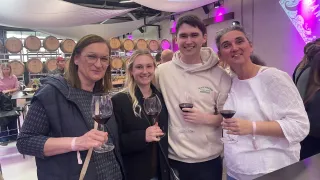 Vier Personen in einem Weingut, lächelnd, mit Weingläsern. Dahinter sind Fässer und eine weiße Decke.