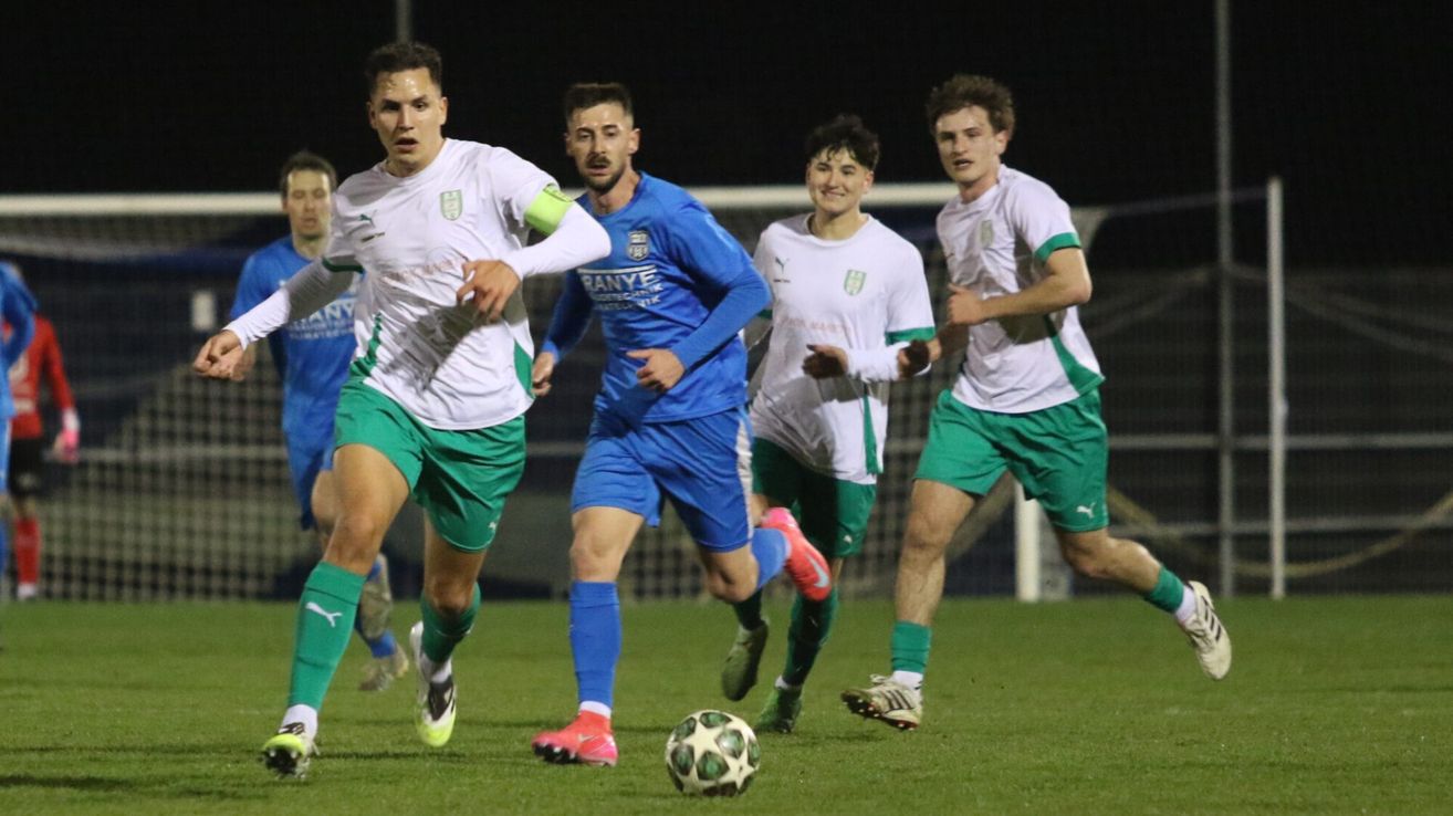 Eine Gruppe von Fußballspielern in grünen und weißen Trikots läuft auf dem Feld und jagt dem Ball hinterher. Der Spieler in Blau versucht, den Ball zu blockieren.