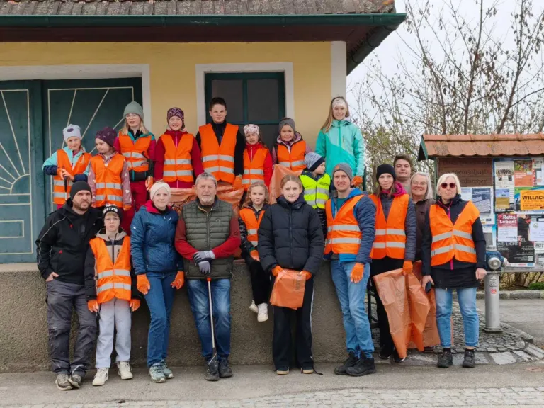 Eine Gruppe von Menschen in orangefarbenen Sicherheitswesten und Handschuhen, die vor einem Gebäude für ein Foto posieren. Einige halten orange Beutel. Dahinter ein Gebäude mit Glasfenstern und einem Baum.