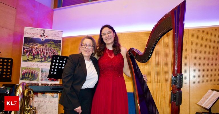 Two women pose with a harp, smiling and looking at the camera, likely at a music event. One wears a red dress and glasses, the other a black suit and white shirt.