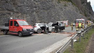 Rettungsfahrzeuge umgeben einen Autounfall am Straßenrand. Ein Auto hat einen beschädigten Motorraum und offene Türen. Helfer in Uniformen beurteilen die Szene.
