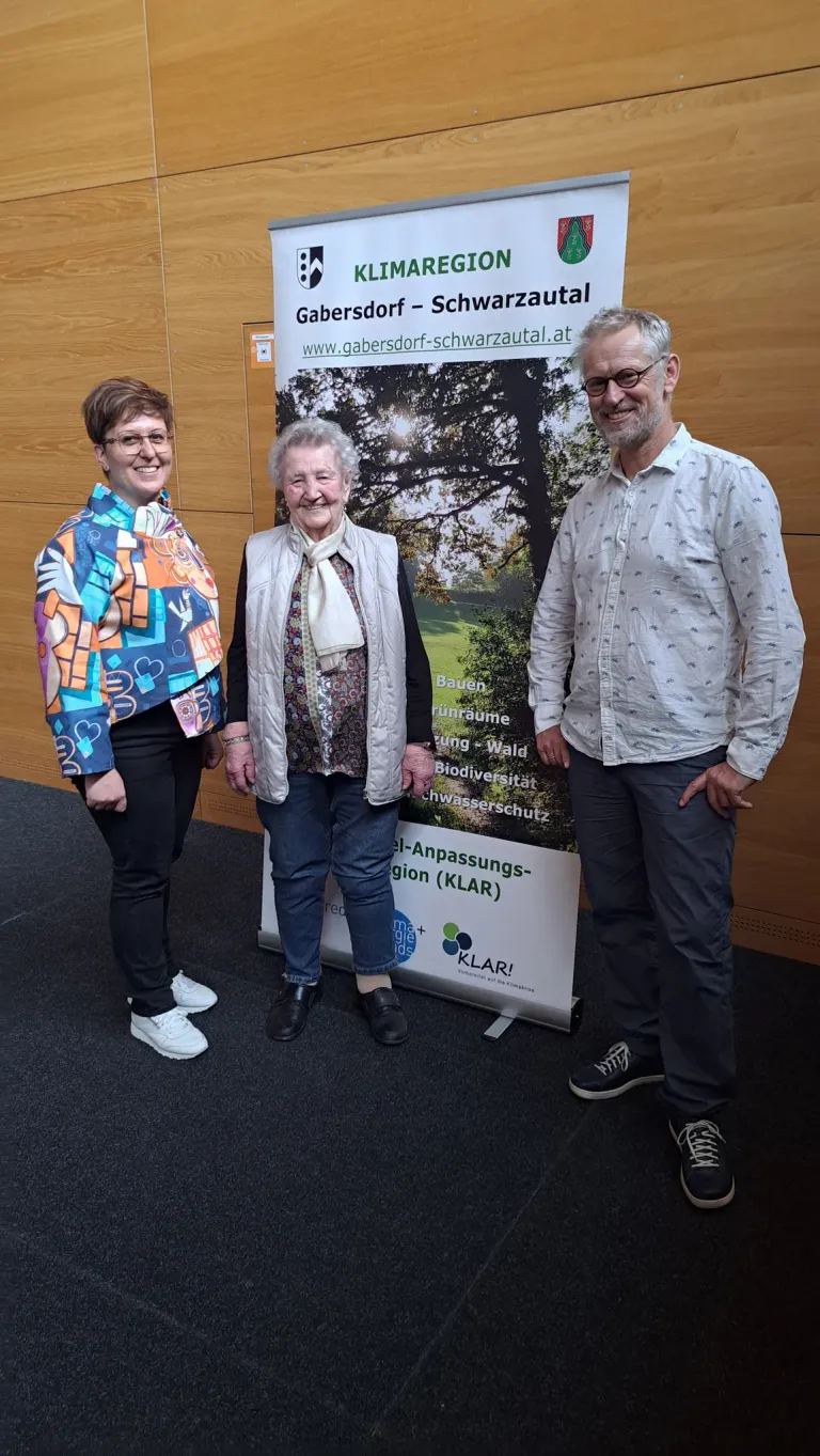 Three people stand next to a banner with a green tree image. They are smiling for the camera. The woman wears colorful clothing, and the man wears glasses.