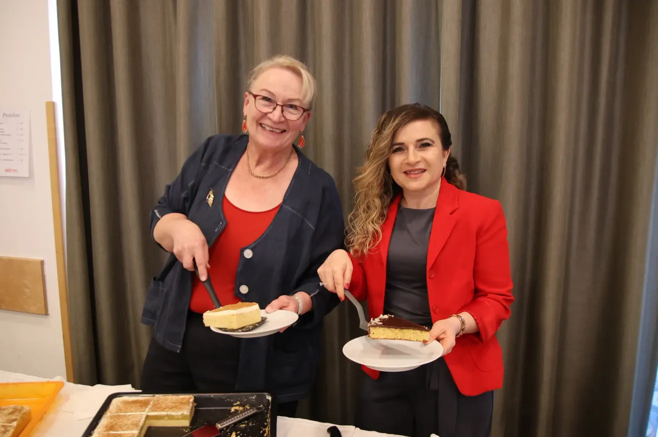 Zwei Frauen in formeller Kleidung, eine mit Messer und die andere mit Gabel, stehen vor einem Tisch mit Kuchenplatten. Die Frau links trägt eine Halskette und eine Brille. Hinter ihnen ist ein Vorhang.