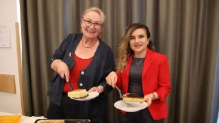 Zwei Frauen in formeller Kleidung, eine mit Messer und die andere mit Gabel, stehen vor einem Tisch mit Kuchenplatten. Die Frau links trägt eine Halskette und eine Brille. Hinter ihnen ist ein Vorhang.