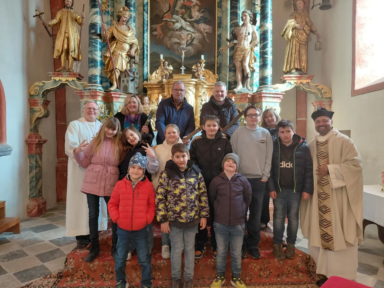 Eine Gruppe von Menschen steht vor einem Kirchenaltar, einige halten Gitarren. Statuen und religiöse Gemälde schmücken den Hintergrund.