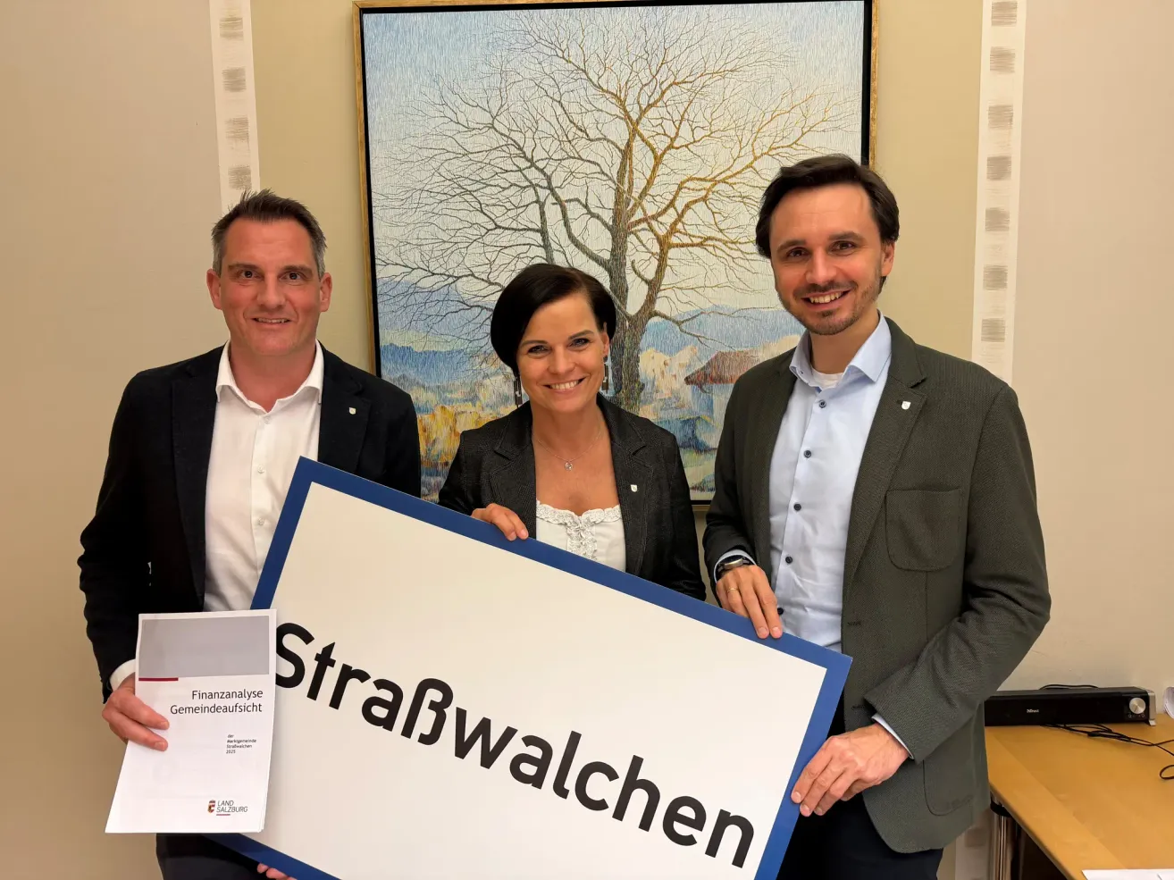 Drei Personen stehen zusammen und halten ein Schild mit der Aufschrift StraBwalchen. Die Frau und ein Mann lächeln, während der Mann auf der linken Seite einen Finanzbericht in der Hand hält.