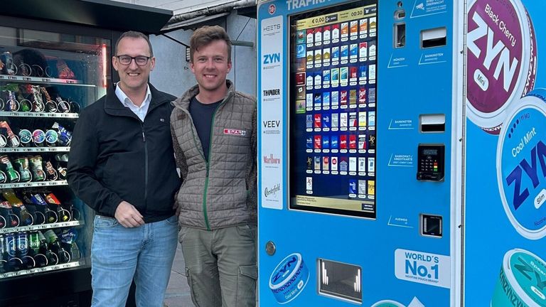 Zwei Männer stehen neben einem blauen Automaten, der verschiedene Zigarettenmarken und ein 'WORLD NO NICOTINE POUCH' Schild zeigt. Ein Mann trägt eine Jacke mit 'SPAR' Schriftzug, und sie lächeln beide für die Kamera.
