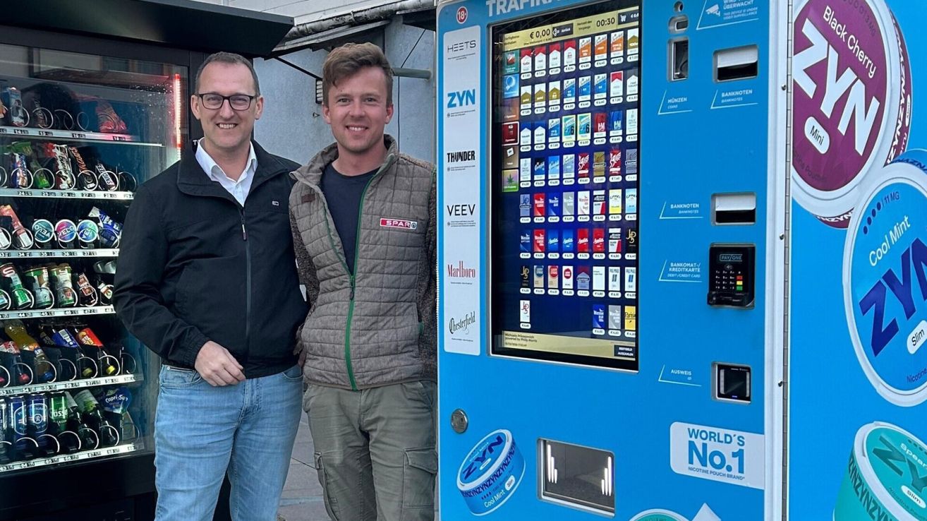 Zwei Männer stehen neben einem blauen Automaten, der verschiedene Zigarettenmarken und ein 'WORLD NO NICOTINE POUCH' Schild zeigt. Ein Mann trägt eine Jacke mit 'SPAR' Schriftzug, und sie lächeln beide für die Kamera.