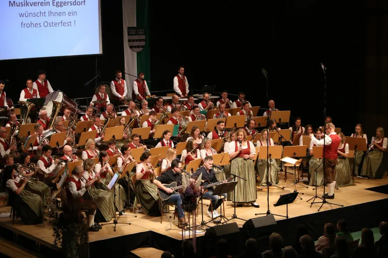 Ein großes Orchester spielt auf der Bühne mit Blech- und Streichinstrumenten, begleitet von einigen Sängern. Das Publikum sitzt im Vordergrund.