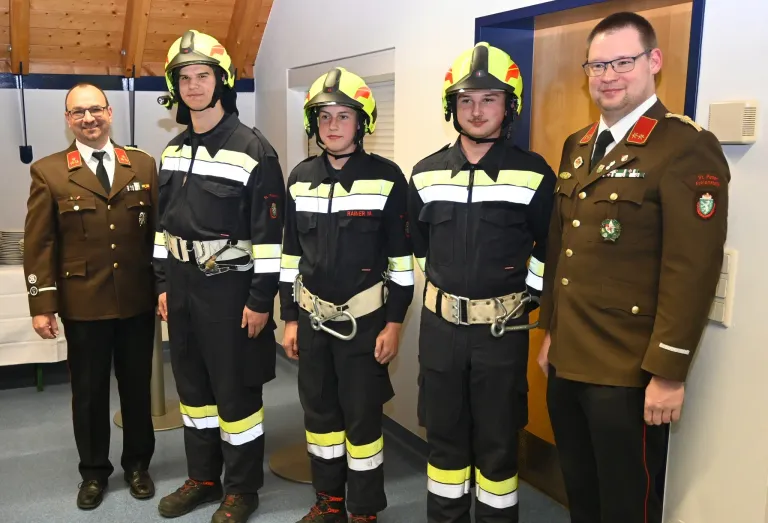 Vier Feuerwehrleute in Uniform stehen zusammen mit einem lächelnden Mann in Uniform. Die Feuerwehrleute tragen Helme, Handschuhe und Stiefel.
