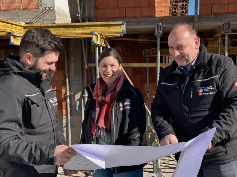 Drei Personen stehen an einer Baustelle. Die beiden Männer lächeln die Frau dazwischen an, die ein Dokument und eine Schleife hält.