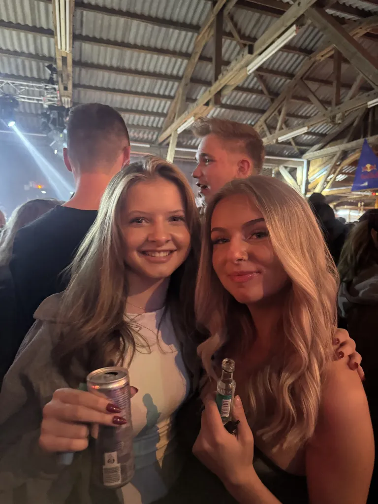 Zwei Frauen lächeln und posieren für ein Foto in einer lebhaften, überfüllten Innenumgebung. Sie halten eine Dose und eine kleine Flasche. Ein Mann ist im Hintergrund sichtbar.