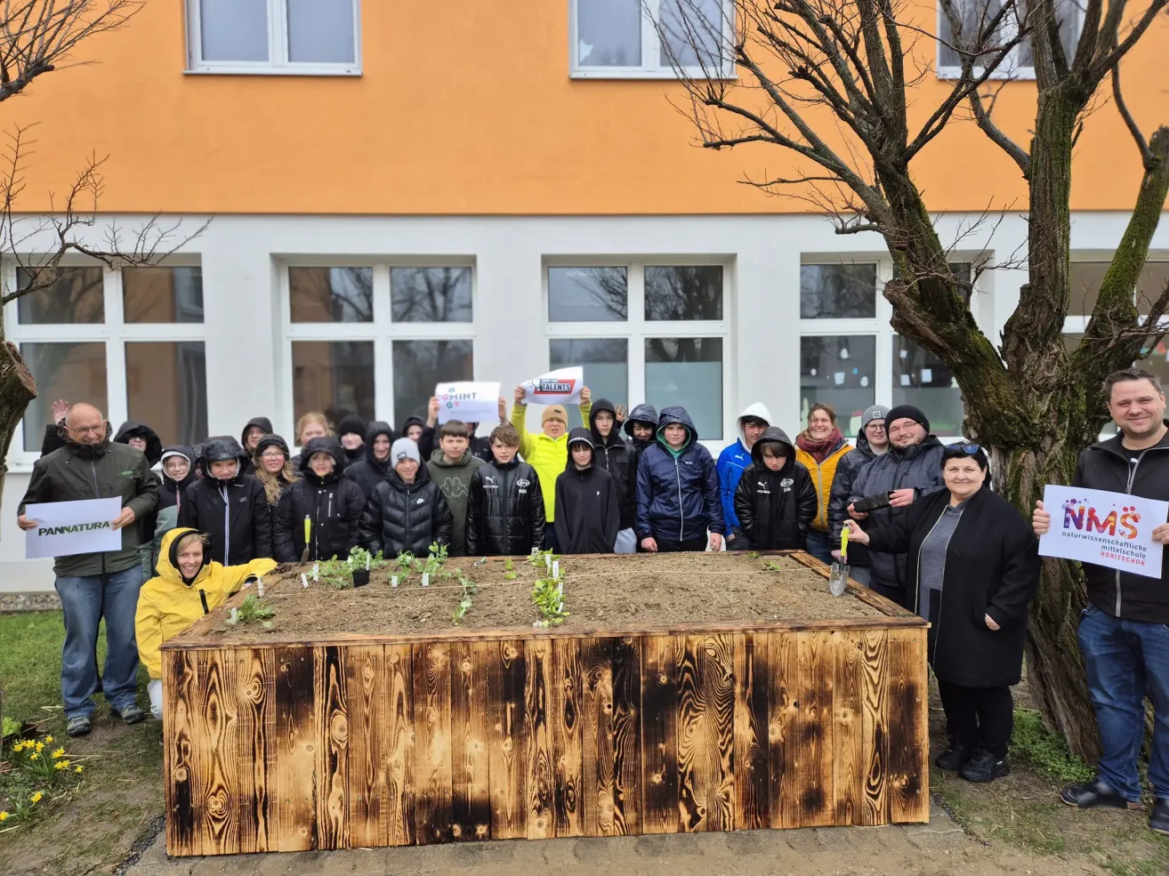 A group of people in winter clothes gather around a wooden planter filled with soil and young plants in front of a building.