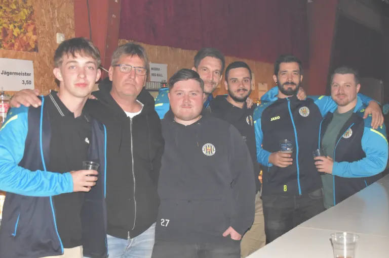 Sechs Männer in identischen Jacken stehen zusammen. Einer trägt eine Brille und hält ein Getränk. Sie scheinen sich bei einer Zusammenkunft zu befinden. Dahinter steht auf einem Schild WOOKA RED BULL.