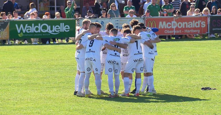 A soccer team huddles on a field, surrounded by spectators. Players wear white uniforms with various sponsor logos. A McDonald's banner is visible in the background.